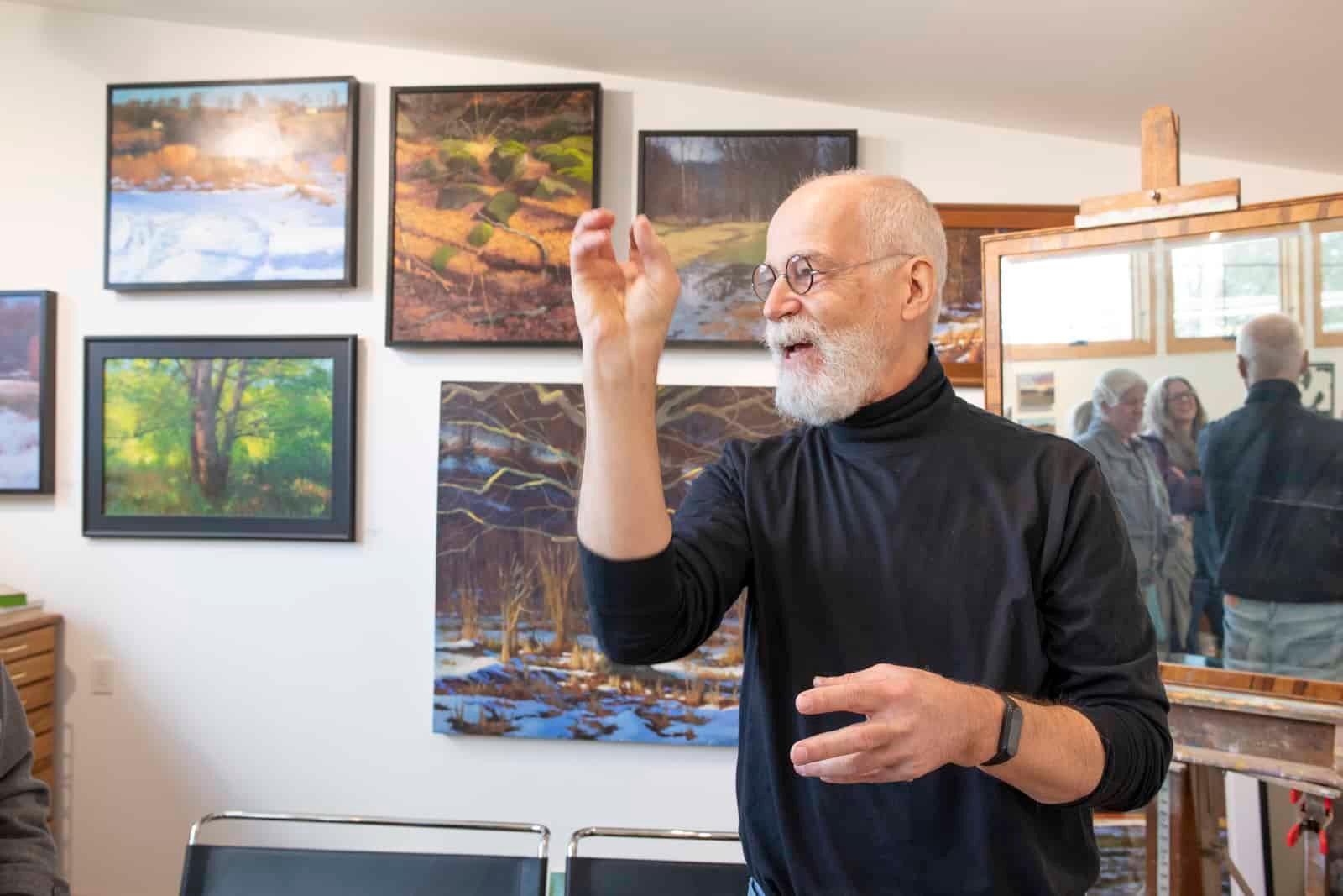 A man wearing a black turtleneck stands in front of his paintings.
