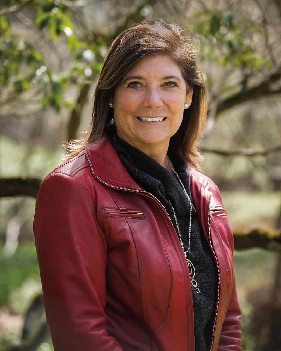 A photo of a woman in a burgundy jacket and brown hair standing outdoors and smiling at the camera.