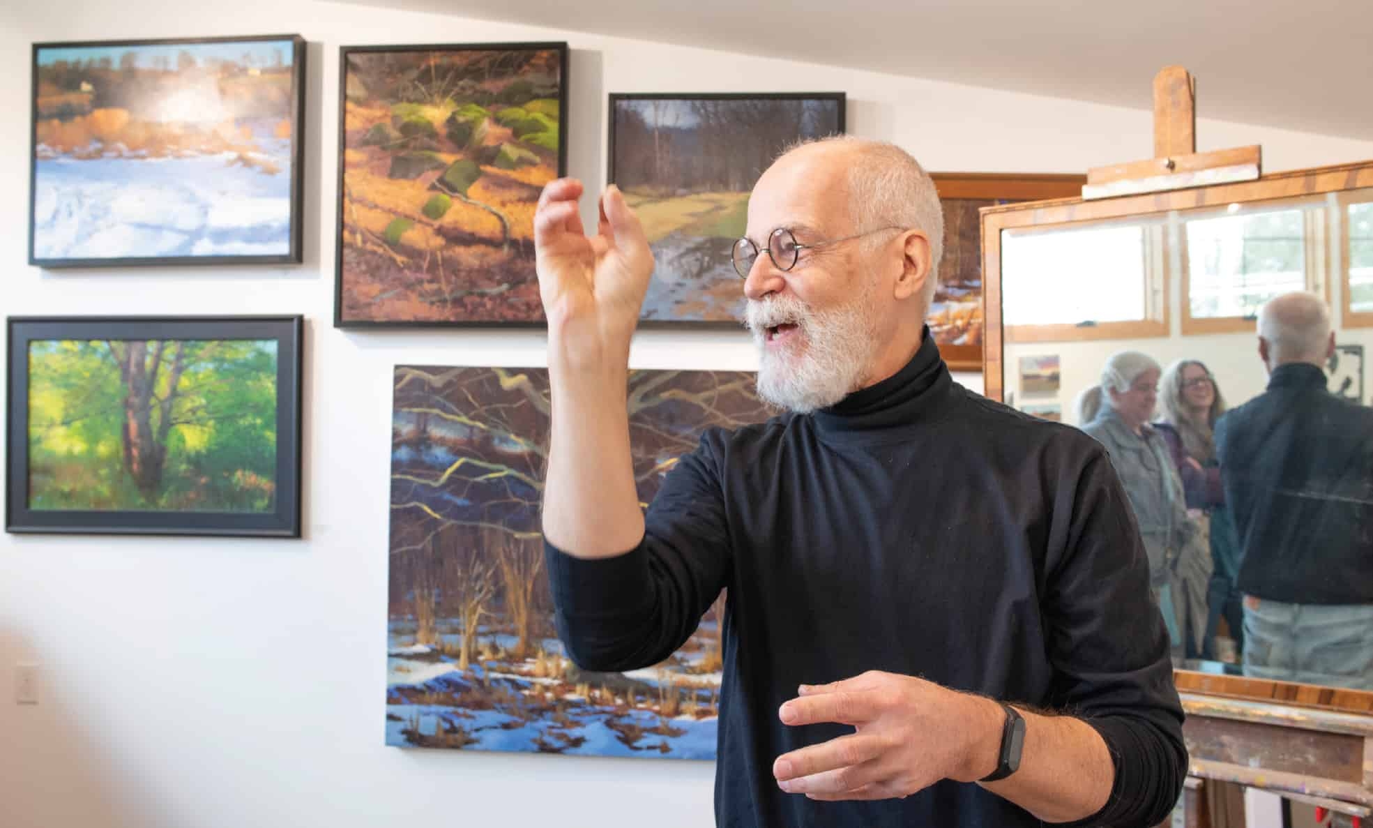 A man wearing a black turtleneck stands in front of his paintings.