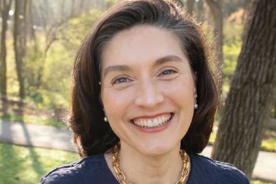A woman stands outdoors smiling at the camera wearing a dark blue sweater