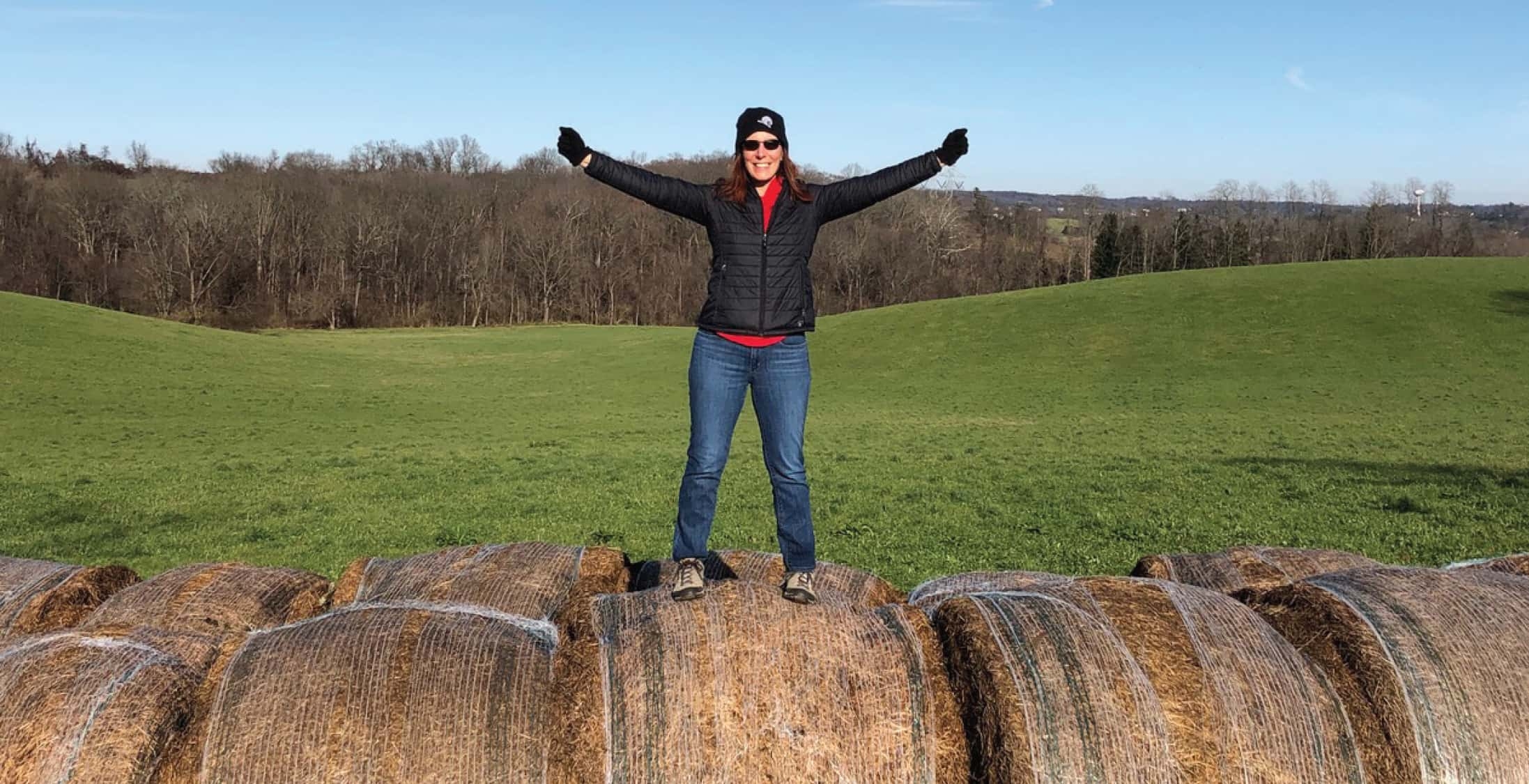A woman in a black coat and hat standing on top of hay bales in a green field.