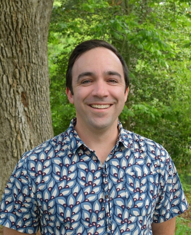 A man in a blue shirt stands outside and smiles at the camera