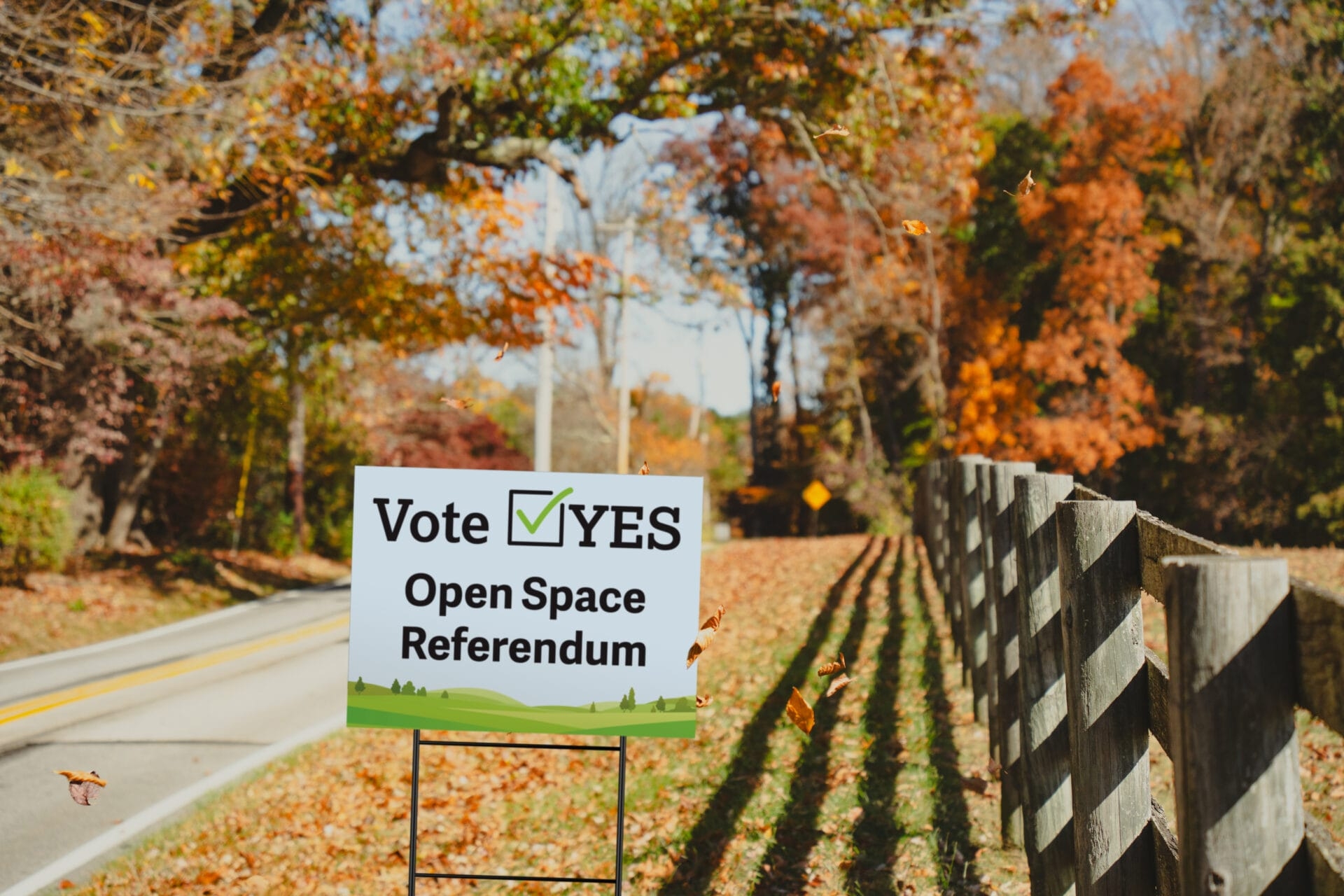 a yard sign that says "Vote Yes Open Space Referendum" along a road with a fence and trees with with orange and red leaves