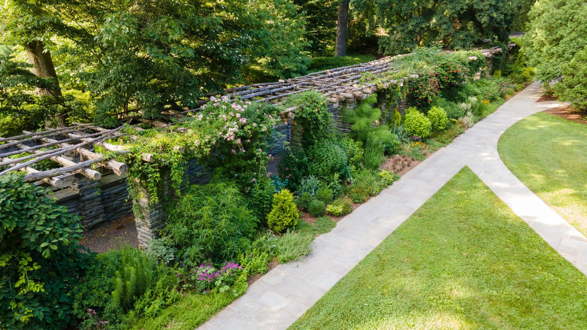 drone photo of large stone pergola with wooden poles on top, lushly planted