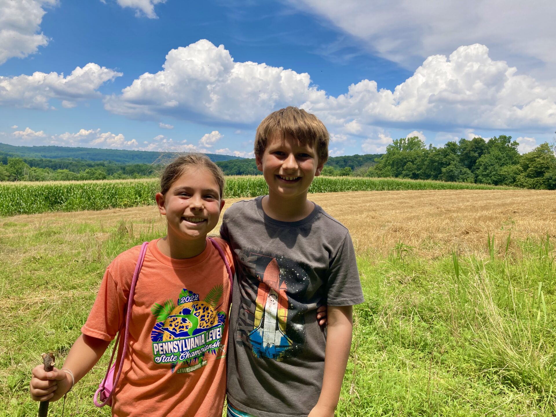 a young boy and girl standing with arms around each other smiling, one with a walking stick, with an agricultural field and blue sky with fluffy white clouds in the background