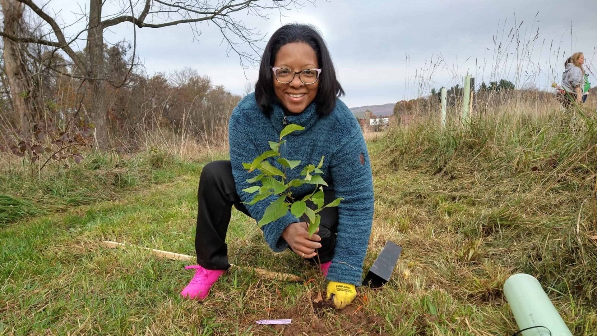Person with glasses smiling and planting a tree sapling into the ground at a nature preserve