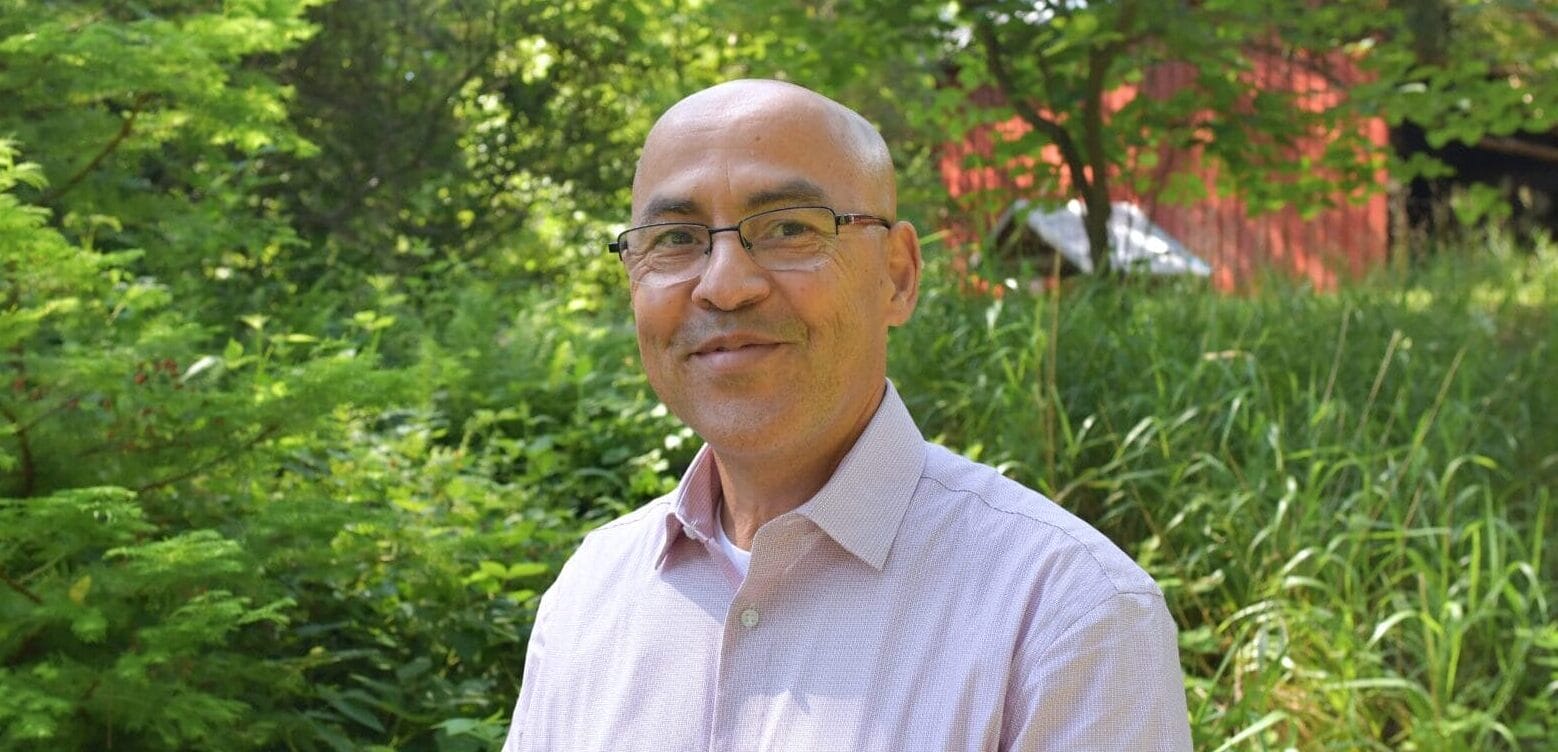 a man with glasses wearing a button-down shirt standing outside with a red barn in the background